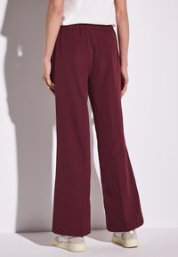 Pantalons larges bordeaux en tissu lisse, dotés d'une taille élastique et de deux poches arrière, associés à des baskets de couleur claire.