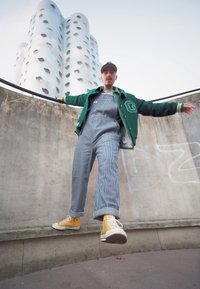 Osoba v zelené varsity bundě přes modré pruhované lacláče, stojící na římsě, s žlutými teniskami Converse a kšiltovkou.