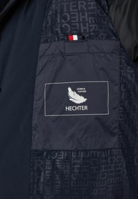 Donkerblauwe jasinterieur met een pocket met een geprinte label, "HECHTER," en een veerlogo. De stof heeft subtiele patroondetails.