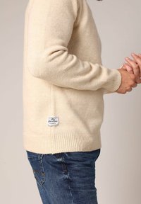 Man in een crèmekleurige gebreide trui met een klein rechthoekig wit label vlakbij de taille en blauwe denim jeans, met de handen voor zich vouwend.