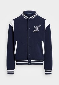 Chaqueta varsity azul marino con acentos blancos, cuello y puños acanalados, con un logo en el frente y dos bolsillos laterales. Textura de tela suave.