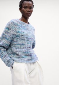 Hellblauer Strickpullover mit strukturiertem Muster und subtilen lila Akzenten. Mit langen Ärmeln und rundem Halsausschnitt, kombiniert mit weißen Hosen.