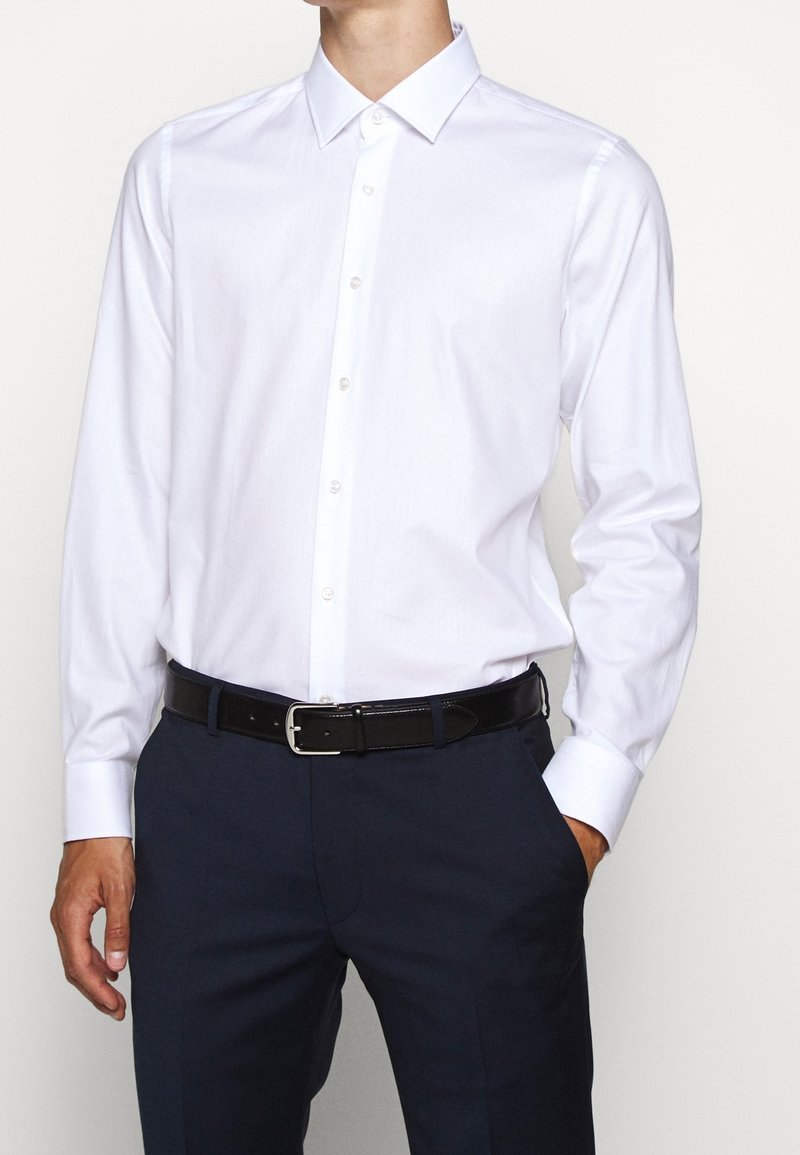 Bílá košile s dlouhým rukávem zapínaná na knoflíky se standardním límečkem, doplněná tmavě modrými kalhotami a černým pásem se stříbrnou sponou.