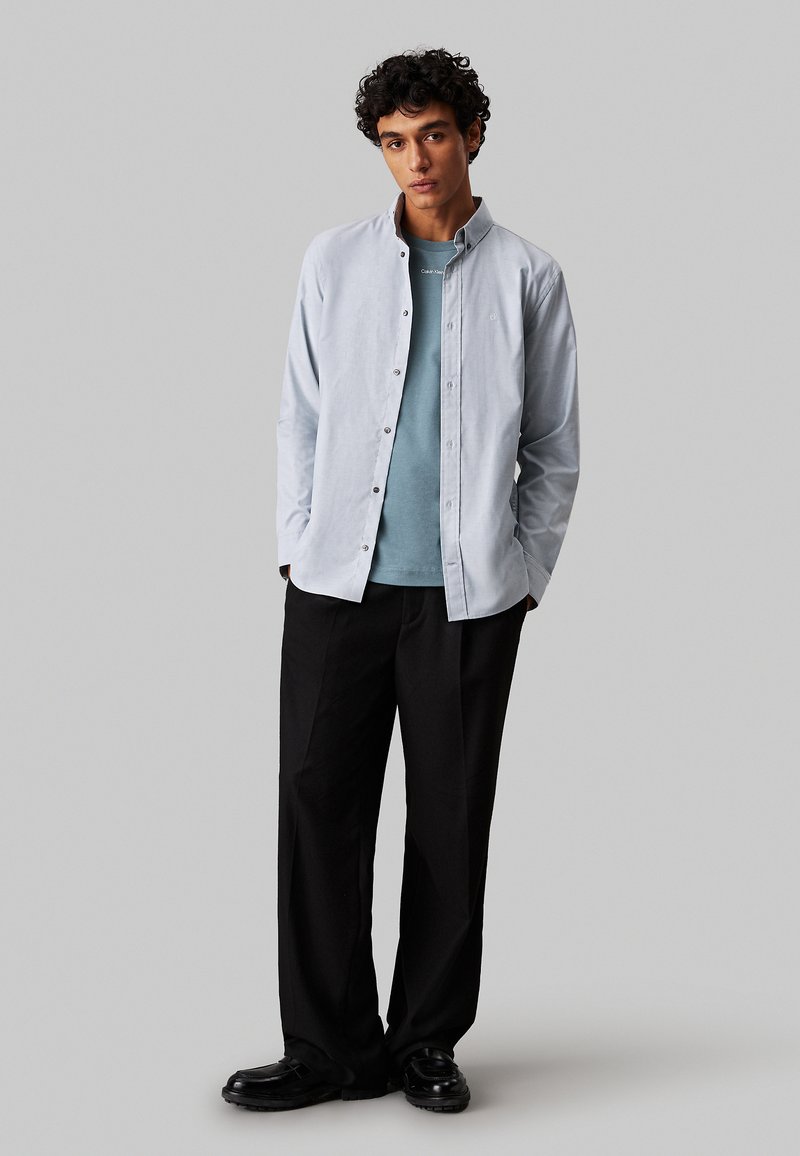 Camisa de botones azul claro, superpuesta sobre una camiseta de color teal, combinada con pantalones anchos negros y zapatos negros. Diseño simple, ajuste casual.