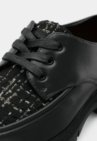 Chaussure noire à lacets avec tige en tissu texturé présentant des fils métalliques et des empiècements en cuir lisse.