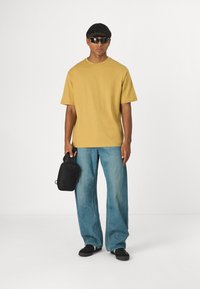 T-shirt kratkih rukava u senfu, široke svijetloplave traperice, nosi malu crnu torbu. Na sunčanim naočalama završava opušteni outfit.
