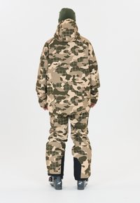 Kamuflage skilædedragt i beige og grøn med et gittermønster, voluminøs design, hætte og forstærkede ankler. Sorte accenter ved manchetterne.