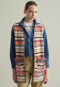 Patterned vest with multicolored stripes, woven texture; features two pockets with tassels in pink and decorative hardware. Layered over denim shirt.