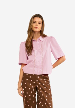 Naine kannab heledat roosat lainelise äärega nööbitavat pluusi, mille juurde on sobitatud pruunid püksid, millel on heledaid roosasid täppe, seistes ühe käega puusal.