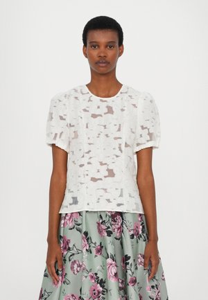 Weiße Bluse mit Puffärmeln aus transparenter Stoff mit strukturierten Blumenmustern; getragen mit einem grünen Blumenrock.