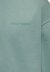 Tela verde claro con el texto "DROPSIZE" en relieve, centrado sobre una superficie de tejido texturizado.