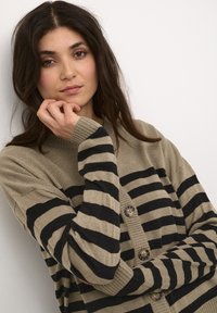 Kootud nööbiga kardigan beežis mustade triipudega. Omab soonikuga varrukaotsi ja lõdva sobivust. Nöörid on siledad ja ümmargused.