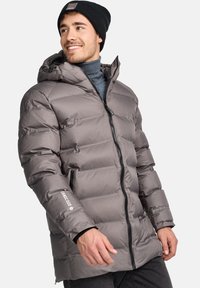 Chaqueta de abrigo gris con capucha, con cierre de cremallera en la parte delantera, textura acolchada y detalle de logo en la manga. Llevada sobre un cuello alto.