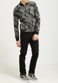 Persona che indossa pantaloni neri, una giacca zip grigia con motivo floreale e sneakers grigie Nike, in piedi contro uno sfondo chiaro e neutro.