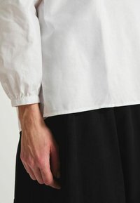 Chemise en coton blanc avec de longues manches froncées, portée sur une jupe noire. La chemise présente un ourlet droit avec un minimum de détails.