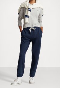 Pantaloni da felpa navy con polsini elasticizzati e vita con cordoncino, abbinati a una maglietta grigia chiara a maniche lunghe e a un maglione beige a maglia appoggiato sulle spalle.