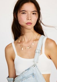 Colier din scoici aurii, cu diverse scoici și farmecuri de culori deschise. Modelul poartă un tricou alb și salopetă din denim, pe un fundal simplu.