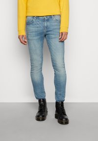 Lichtblauwe slim-fit jeans van denim, gecombineerd met zwarte veterschoenen. De persoon draagt een gele langslijfshirt.