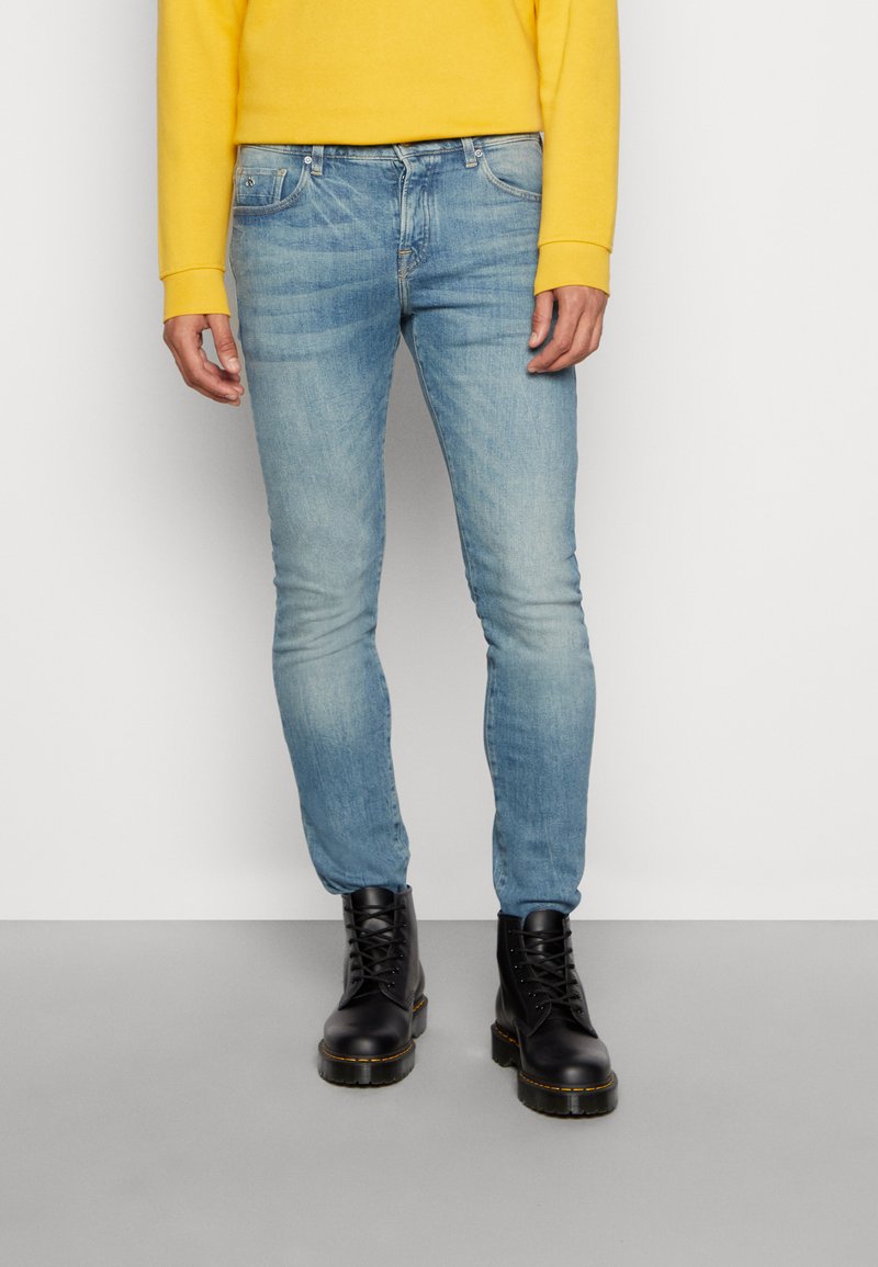 Lichtblauwe slim-fit jeans van denim, gecombineerd met zwarte veterschoenen. De persoon draagt een gele langslijfshirt.