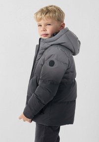 Chaqueta acolchada gris con capucha, con cierre frontal de cremallera, puños elásticos y un parche circular con logo en el brazo izquierdo. Textura suave y diseño cálido.