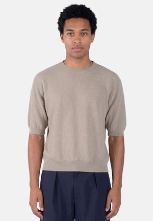 Uomo con capelli corti e ricci che indossa un maglione beige a maniche corte e pantaloni blu scuro, in piedi davanti a uno sfondo semplice.