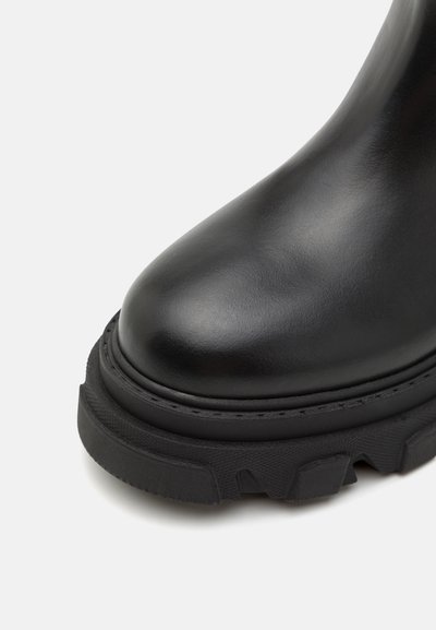 Črne usnjene gležnjarje z zaobljenim nosom, debelim črnim gumijastim podplatom z teksturiranimi detajli in vidno šivano obrobo.
