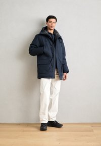 Navy insulated coat with a hood, front pockets, and button closure, paired with white pants and black sneakers on a wooden floor.
