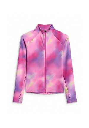 Młoda dziewczyna stoi ubrana w różowo-fioletową kurtkę na zamek z efektem tie-dye, czarne spodnie i czarne trampki na jednolitym szarym tle.