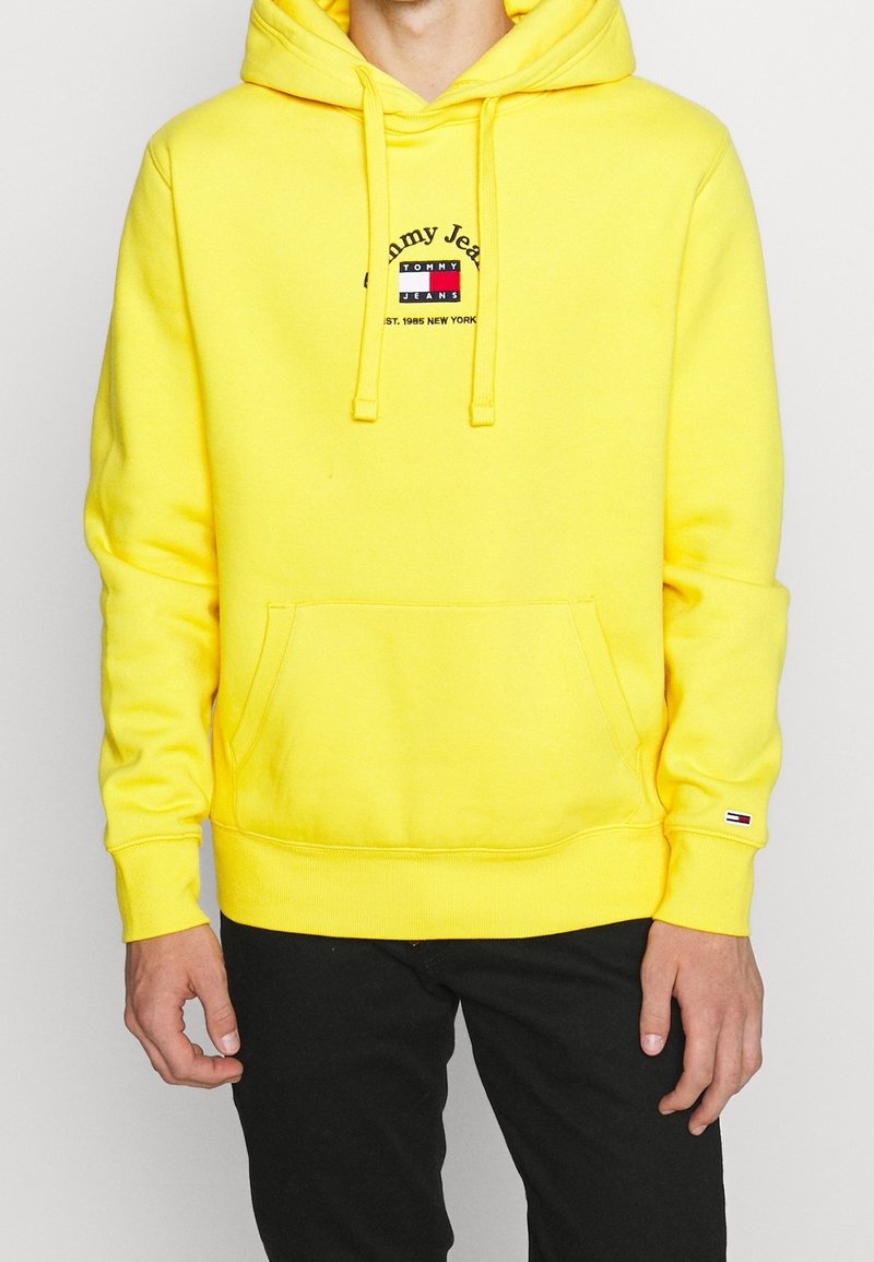 Homme portant un sweat à capuche jaune vif Tommy Jeans avec poche avant et pantalon noir, debout devant un fond blanc.