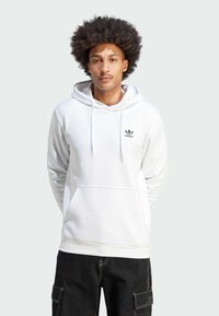 Felpa bianca in cotone con cappuccio e cordini, tasca frontale a marsupio e logo Adidas nero sul petto. I pantaloni cargo neri completano l'outfit.