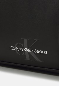 Crna kožna torba s sjajnom površinom, urezanim 'CK' logom i bijelim tekstom koji glasi 'Calvin Klein Jeans' na prednjoj strani.