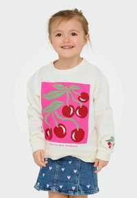 Sweatshirt léger crème avec graphique de cerises roses, feuilles vertes et texte. Associé à une jupe en denim ornée de motifs en forme de cœur.