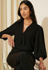 Blusa nera con dettaglio della scollatura a cravatta e maniche a palloncino, realizzata in un tessuto liscio. Il modello indossa pantaloni neri e siede su una sedia trasparente.