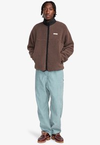 Giacca in pile marrone con colletto nero, chiusura a zip e tasca frontale; abbinata a jeans azzurri chiari e calzature marroni.