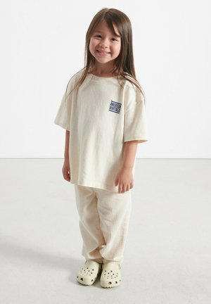 Beeżikas toonitud suur t-särk, millel on sinine "American Vintage" logo, kombineeritud heledate triibuliste pükstega ja kreemjate klotskontsadega. Siledad, pehmed materjalid.