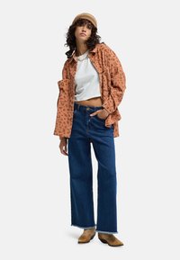 Camicia a righe arancione ruggine, top corto bianco, jeans larghi blu e stivali marroni. Tessuto strutturato e design casual.