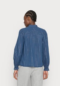 Blusa azul riscada com mangas longas, tecido texturado, colarinho em babados e punhos elásticos, combinada com calças cinza claro.