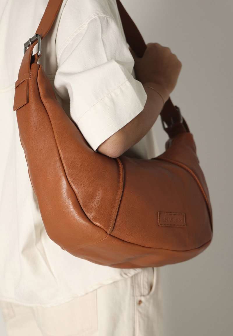 Borsa a tracolla in pelle marrone con una forma morbida e arrotondata. Presenta una singola tracolla e un logo inciso discreto. Texture liscia e linee pulite.