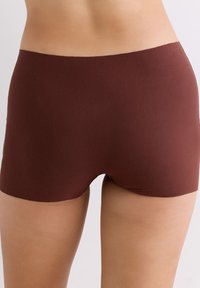 Close-up van de onderkant van het lichaam van een persoon die naadloze, donkerbruine boyshort-onderbroek draagt tegen een eenvoudige lichte achtergrond.