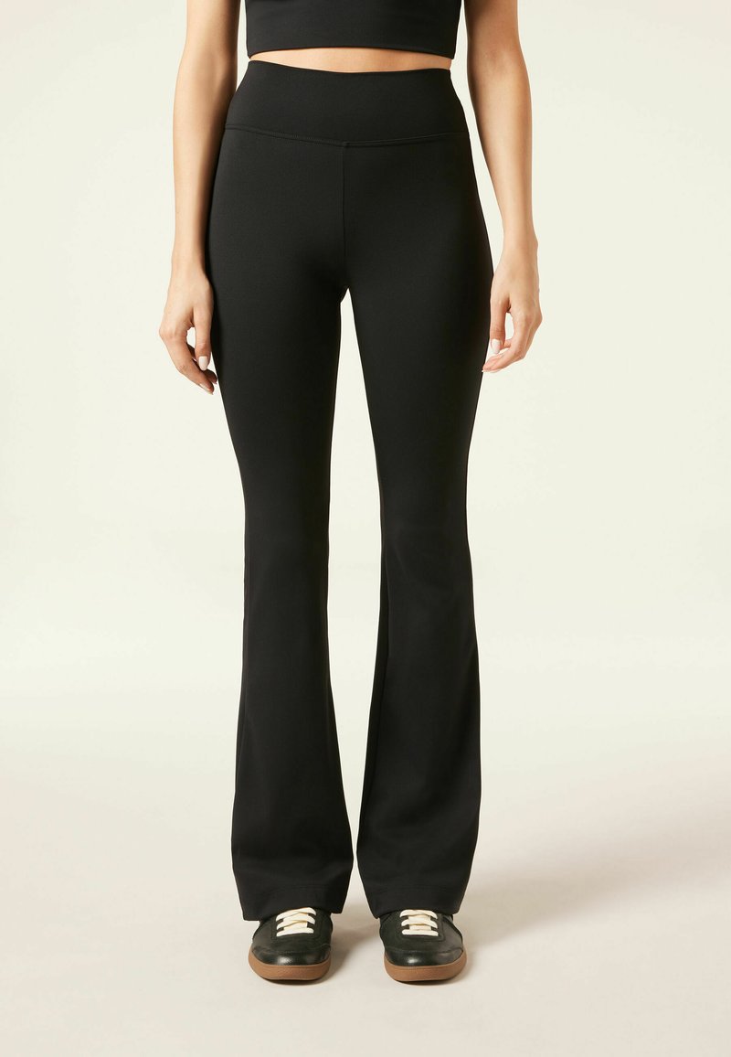 Femme portant un pantalon noir évasé taille haute et des baskets noires avec des lacets blancs, se tenant contre un fond clair uni.