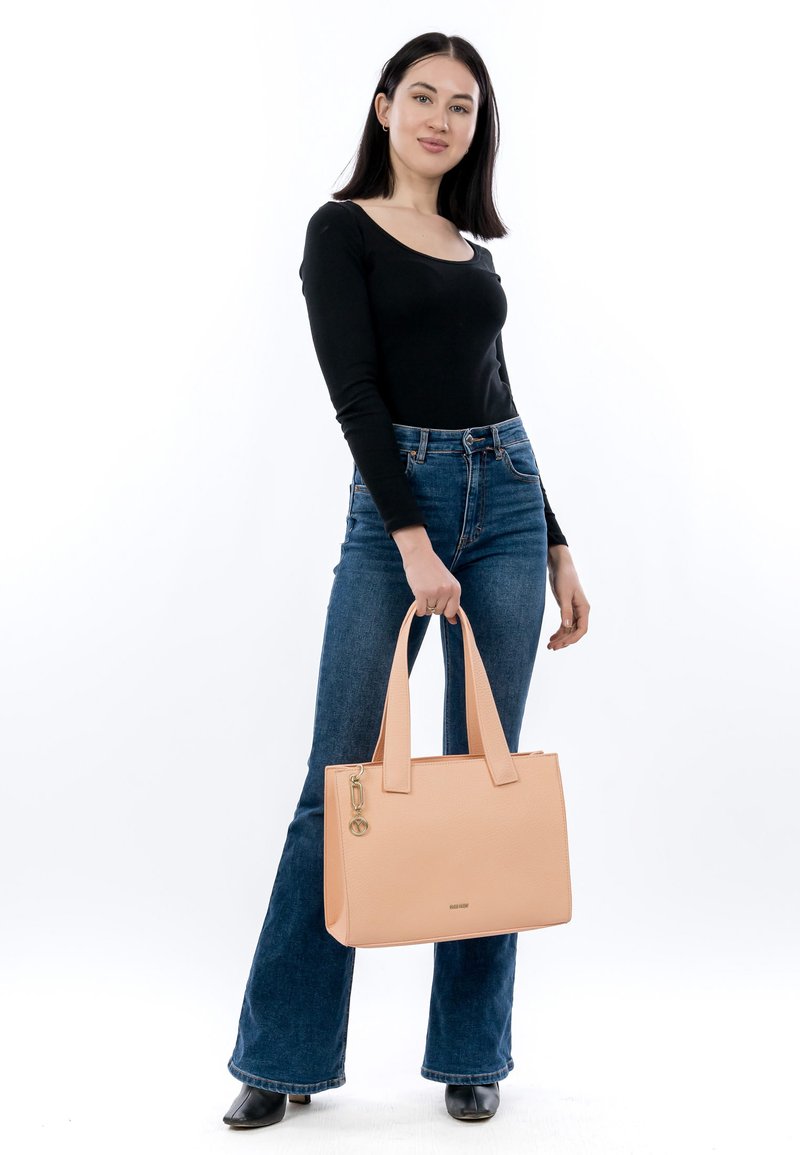 Lyserød læder tote taske med struktureret design, dobbelte top håndtag og et charms detalje. Model holder tasken mens hun har en sort top og flagrende jeans på.