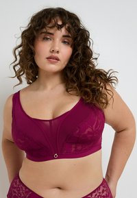 Donna dai capelli ricci che indossa un reggiseno in pizzo color magenta scuro e un paio di mutande coordinate su uno sfondo chiaro e semplice.