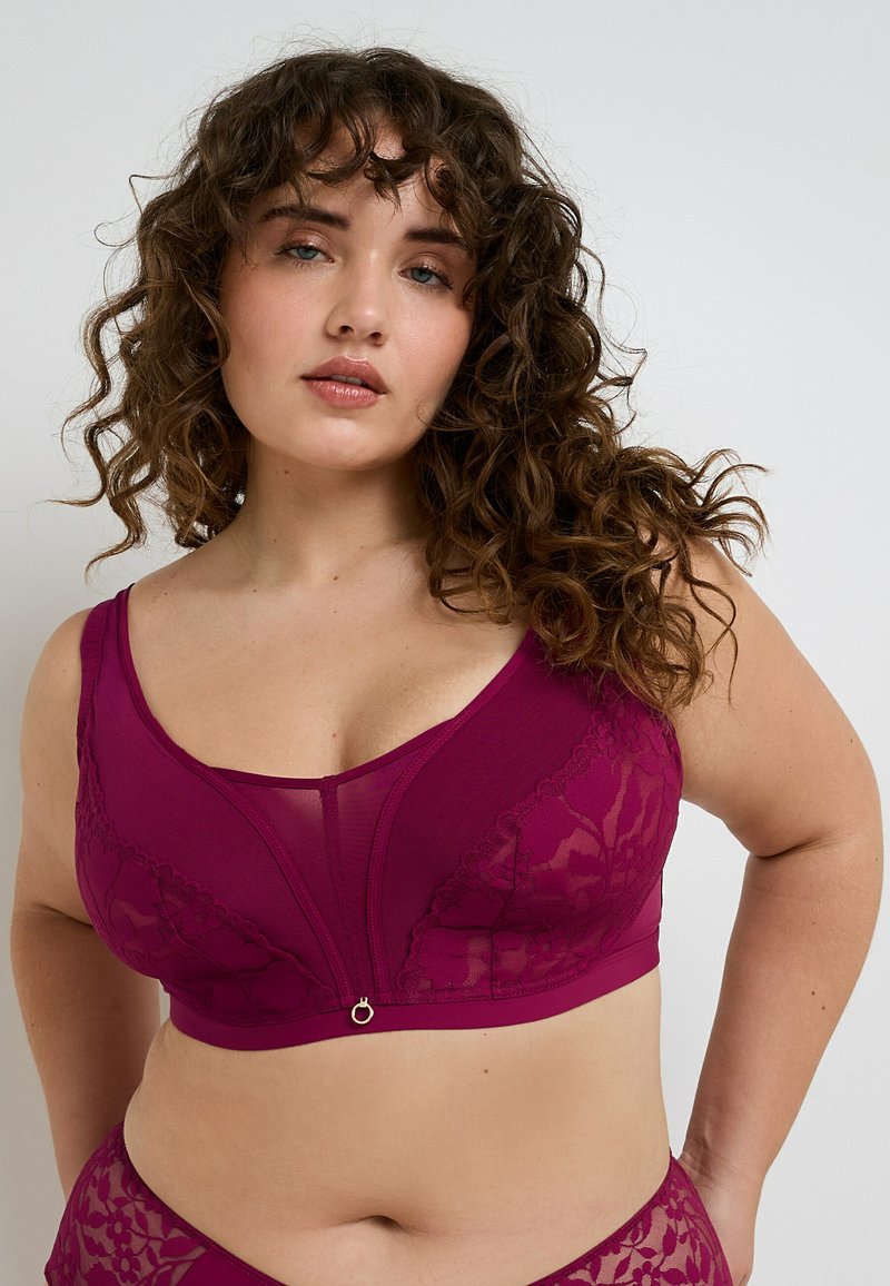Donna dai capelli ricci che indossa un reggiseno in pizzo color magenta scuro e un paio di mutande coordinate su uno sfondo chiaro e semplice.