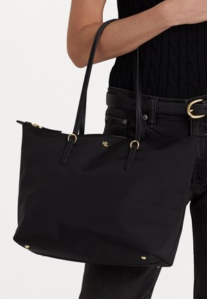 Personne tenant un grand sac fourre-tout noir avec double poignée et détail de fermeture éclair dorée, portant des vêtements noirs et une ceinture avec boucle dorée.