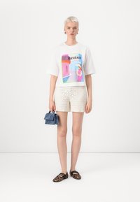 White t-shirt with a colorful graphic print, paired with beige textured shorts featuring a floral pattern. Accessory: small blue handbag.