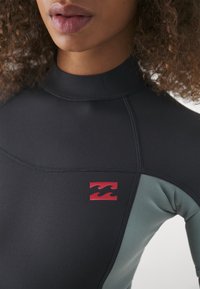 Combinaison en néoprène noire avec des accents turquoise clair, col montant et coutures plates. Présente un logo rouge avec deux formes de vagues sur le côté. Texture douce.