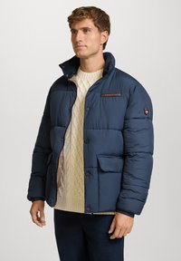 Giubbotto puffer blu navy con colletto alto, chiusura a bottoni, tasca frontale e dettaglio logo. Materiale imbottito con una finitura liscia.