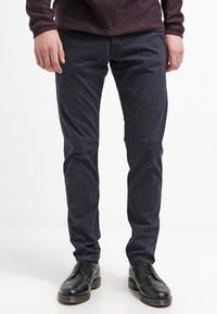 Muž v tmavě modrých slim-fit kalhotách, černých kožených formálních botách a vínově červené vzorované košili s dlouhým rukávem stojí na bílém pozadí.