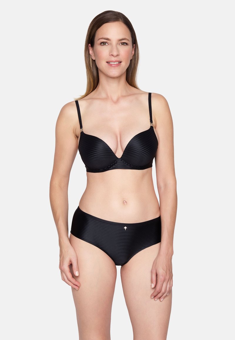 Femme portant un soutien-gorge côtelé noir et un slip taille haute assorti, debout et souriante sur un fond clair uni.