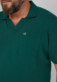Koszulka polo w ciemnozielonym kolorze, wykonana z fakturowanej tkaniny, z dekoltem w serek i kieszonką na piersi z drobnym logo.